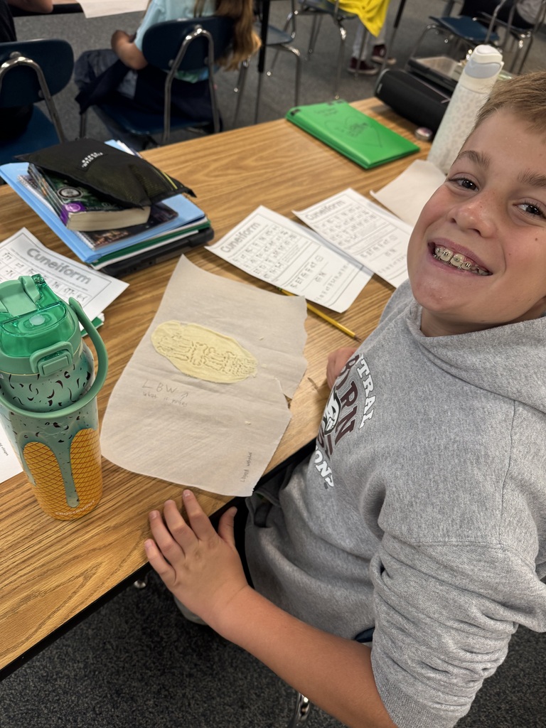 A smiling student at a desk showing off his completed cuneiform clay tablet project next to class materials.
