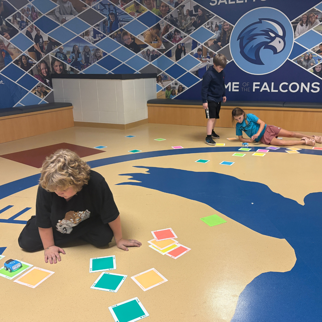 Students sitting on a school floor near a falcon mascot wall, programming small educational robots using colorful tiles