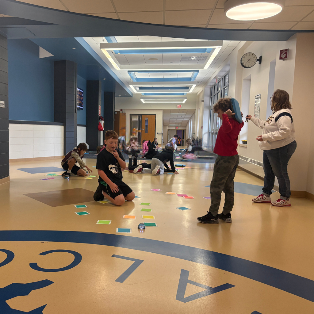 Students in a school hallway using small robots to navigate paths made of colorful coding cards on the floor while a teacher observes.