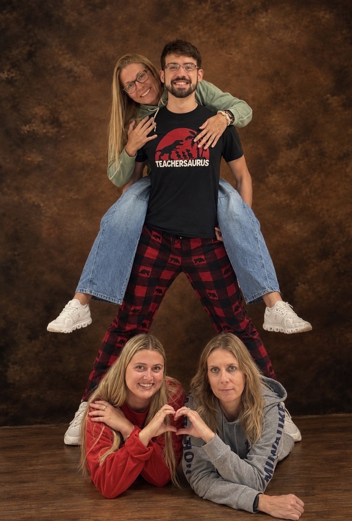 Four school staff members posing playfully together against a studio backdrop, with one piggybacking on another and two lying on the floor below making a heart shape.