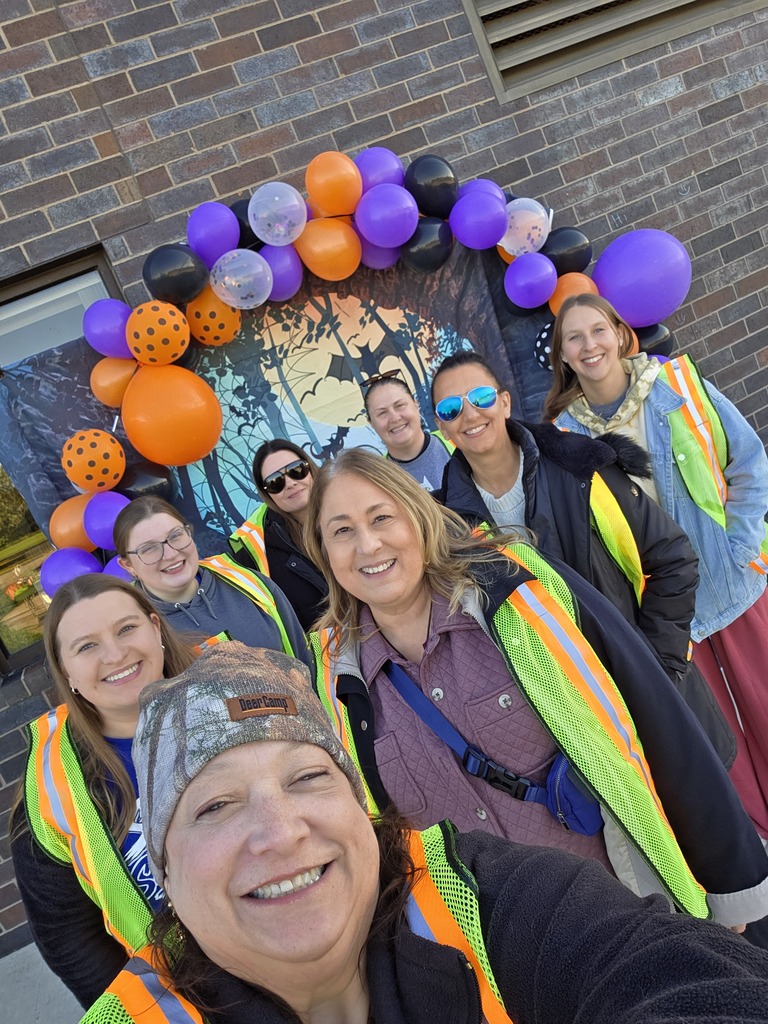 A group selfie of smiling school staff wearing high-visibility safety vests outdoors in front of a Halloween balloon backdrop.