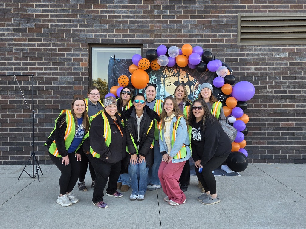 A group of smiling school staff members wearing safety vests posing in front of an orange and purple Halloween balloon arch outside.