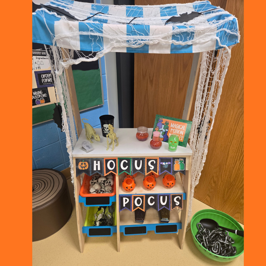 A wooden classroom play station decorated with a "Hocus Pocus" banner, pumpkin buckets, toy skulls, and potion bottles for Halloween.