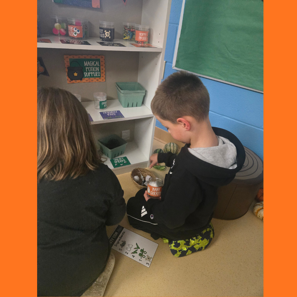 Two young elementary students sitting on the classroom floor sorting toy ingredients at a "Magical Potion Supplies" shelf for a Halloween learning activity.