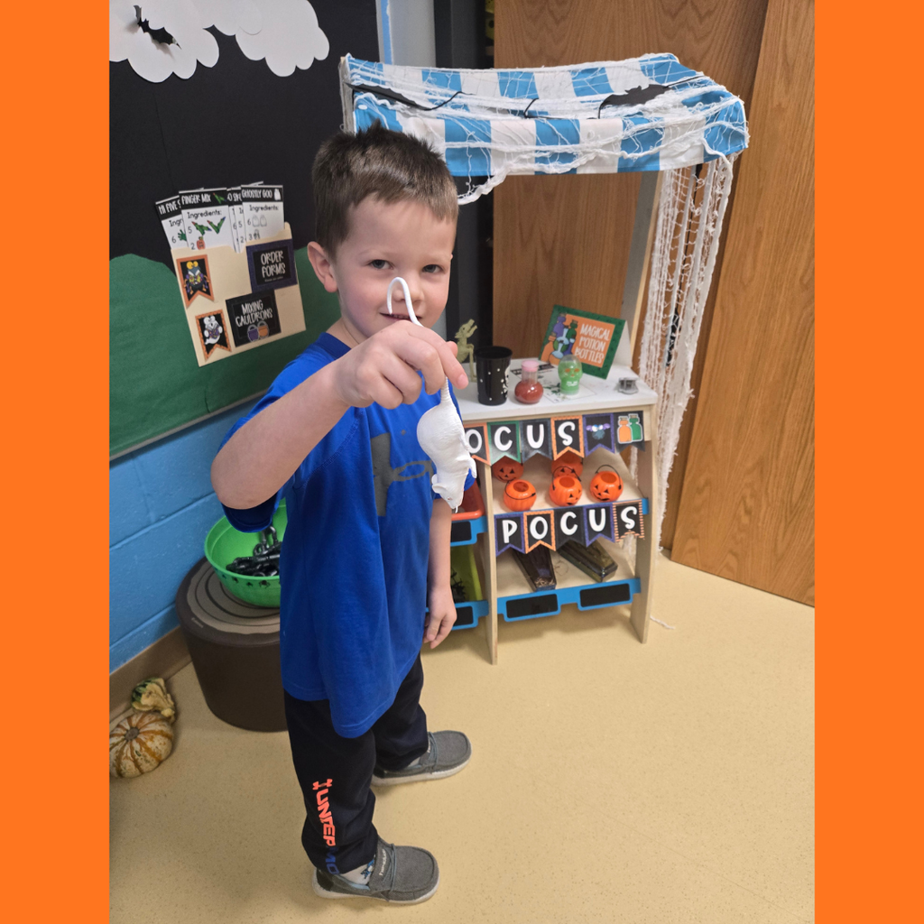 A young boy standing in a classroom proudly holding up a white plastic toy mouse in front of a Halloween play booth.