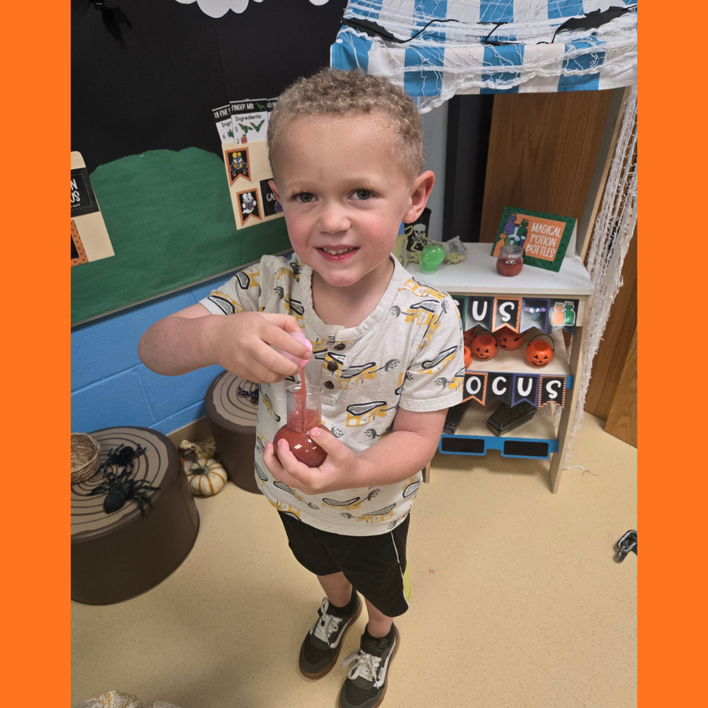 A smiling young student holding a plastic potion bottle during a Halloween classroom activity near a "Hocus Pocus" play stand.