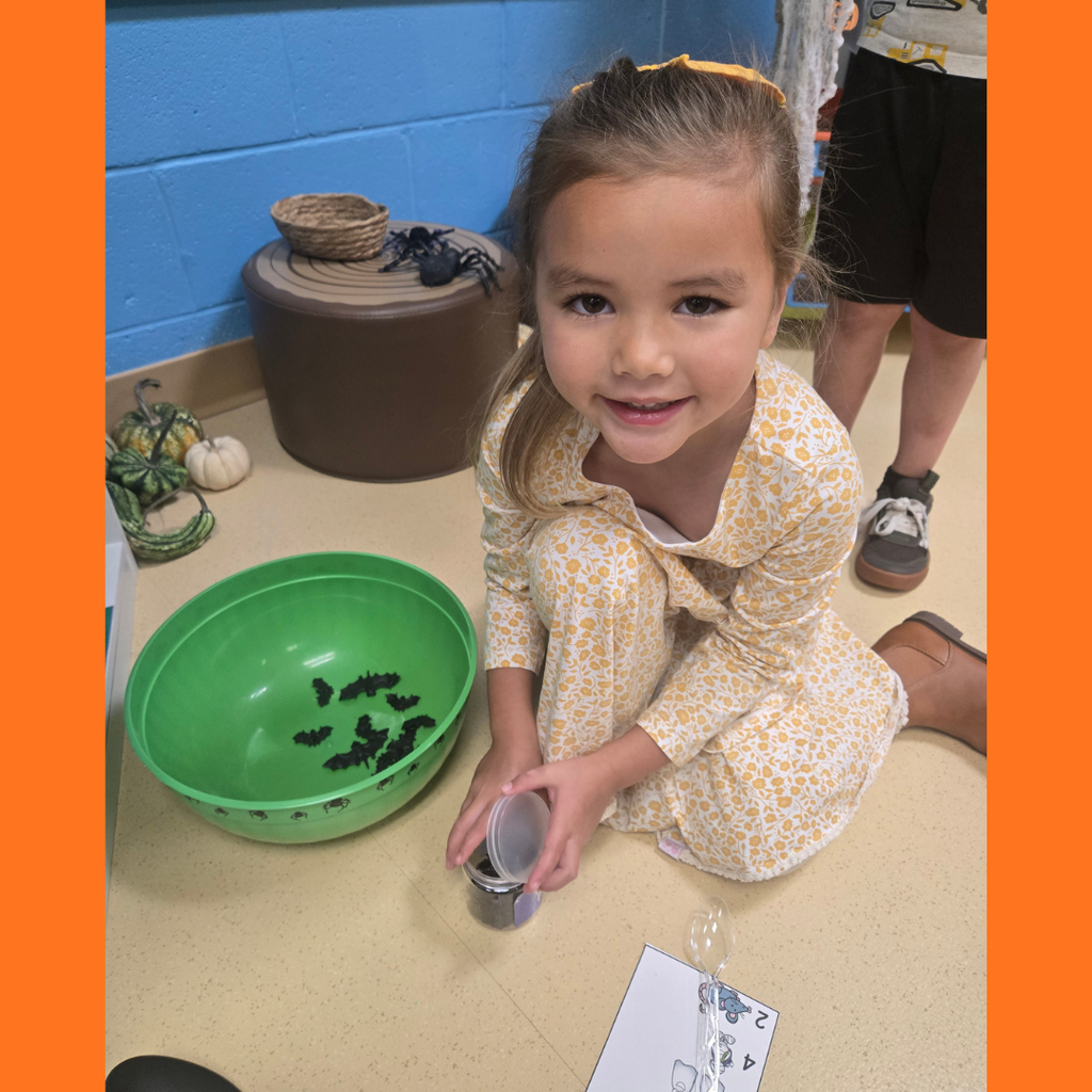 A young girl in a patterned dress smiling while kneeling on the floor next to a green bowl filled with toy bats for a classroom lesson.