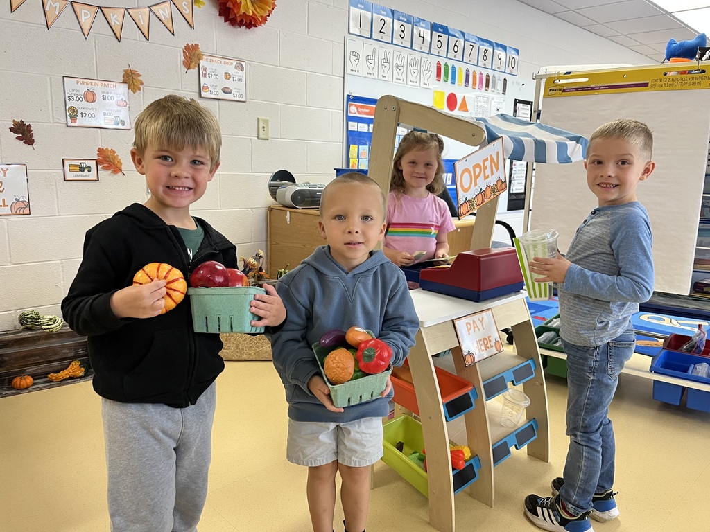Four young students smiling during a pretend farmers market learning activity, holding baskets of play produce near a wooden market stand.