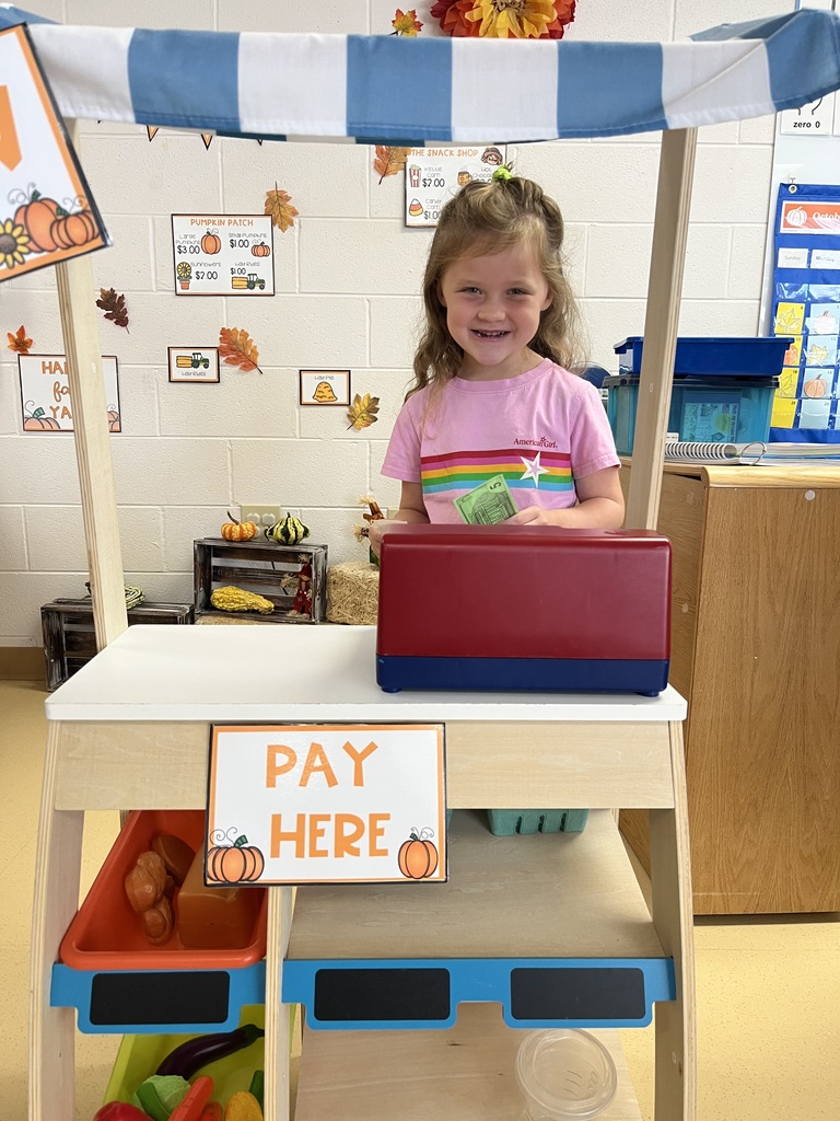 A smiling young student standing behind a wooden play market stand with a toy cash register and a "PAY HERE" sign.