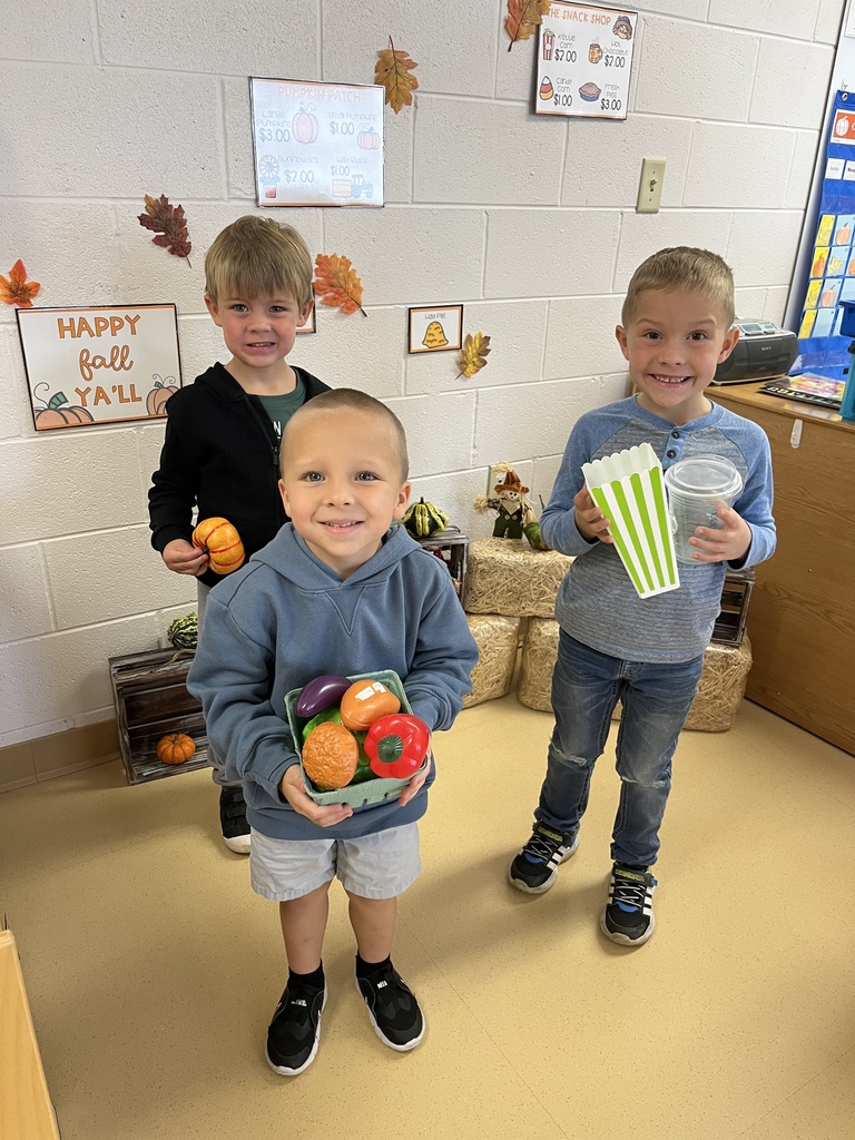 Three young students posing together holding baskets of toy produce and a pretend popcorn container during a classroom lesson.