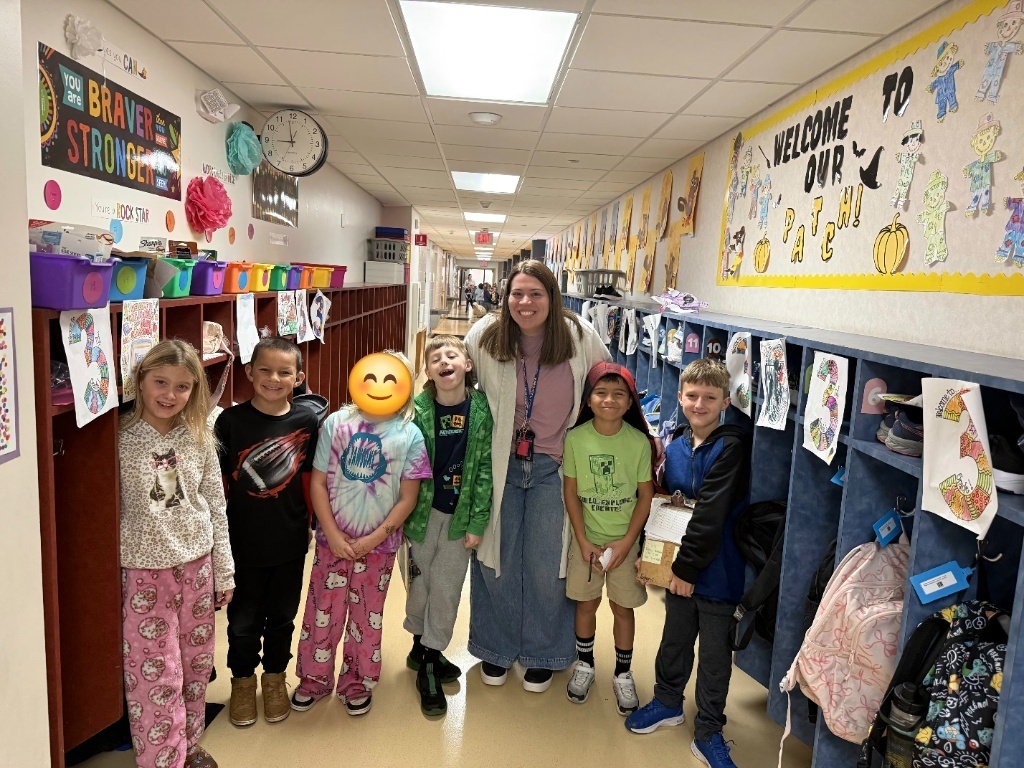 A smiling teacher standing in a school hallway with six elementary students in front of student cubbies and decorated bulletin boards.