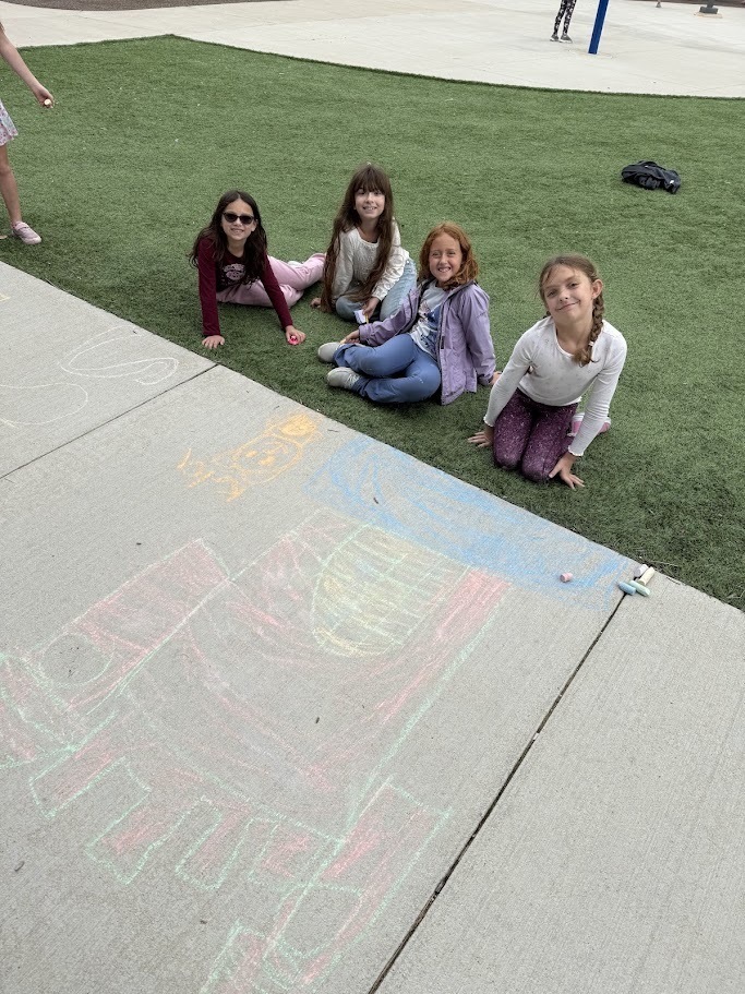 Four elementary school girls sitting together on green turf outside next to sidewalk chalk drawings.