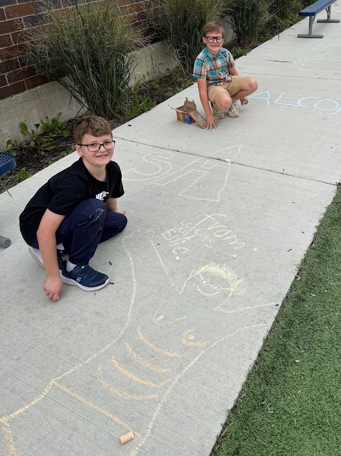 Two young boys smiling while crouching next to sidewalk chalk drawings.