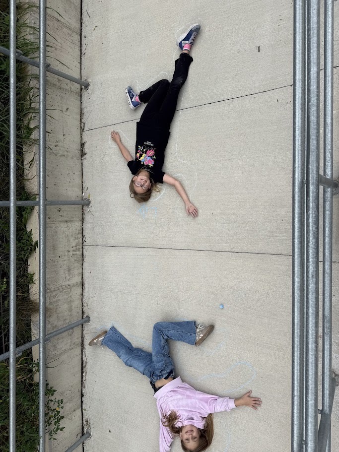 Two elementary students lying on a sidewalk inside drawn chalk body outlines.