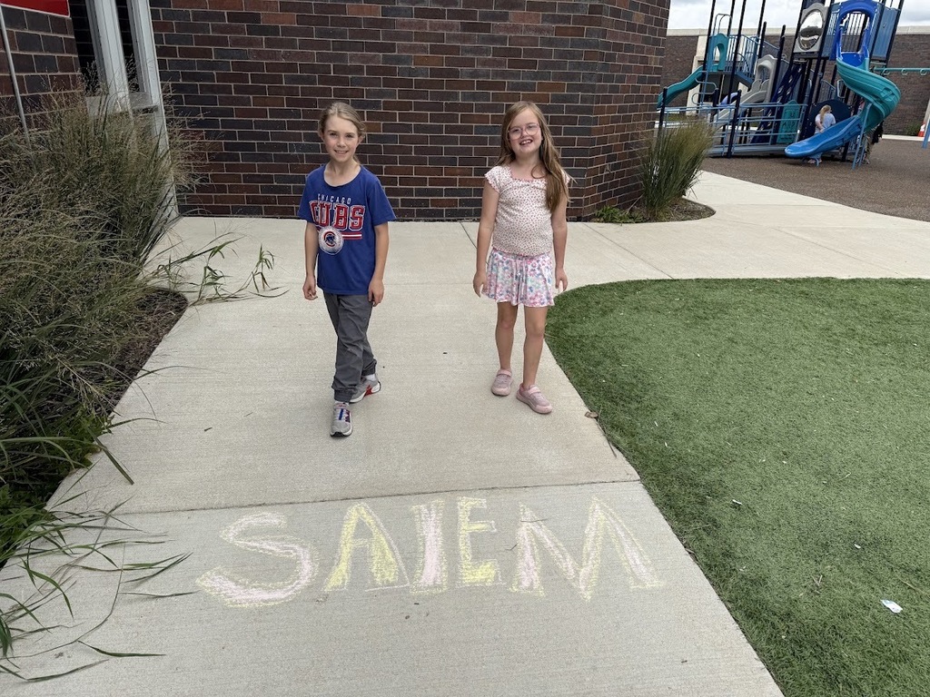 Two elementary school girls standing on a sidewalk near sidewalk chalk art and a playground.