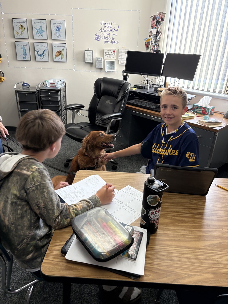 Two students work at a classroom desk; one smiles at the camera while the other pets a golden retriever dog.