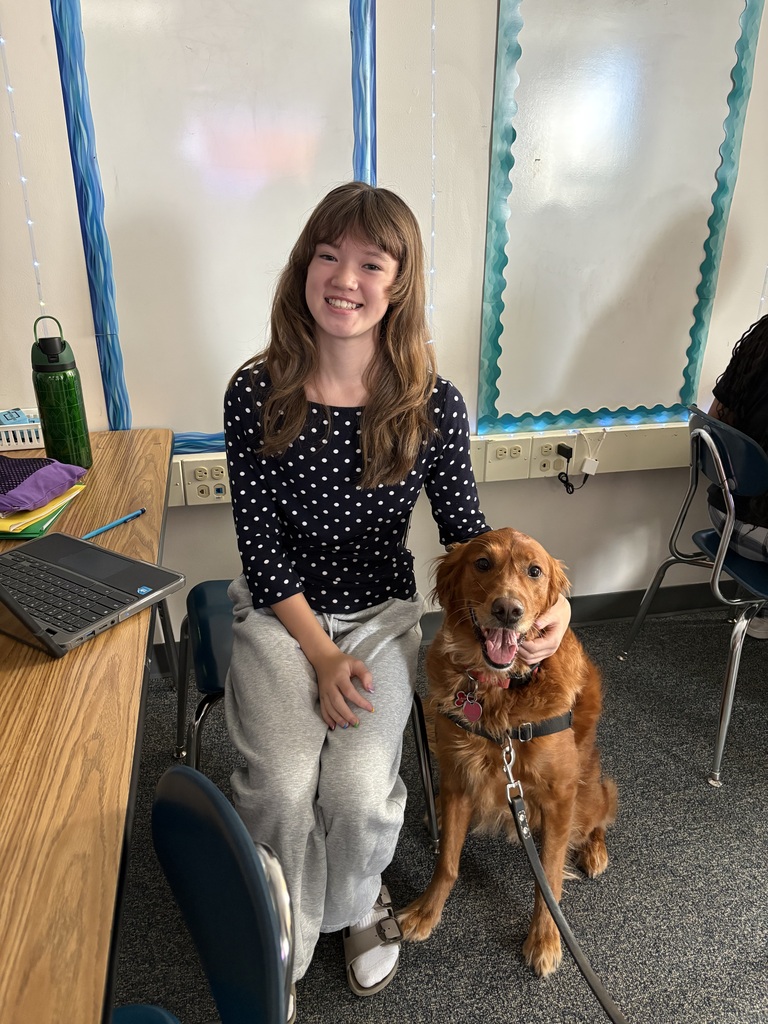 A smiling student poses next to a friendly golden retriever therapy dog in a classroom.