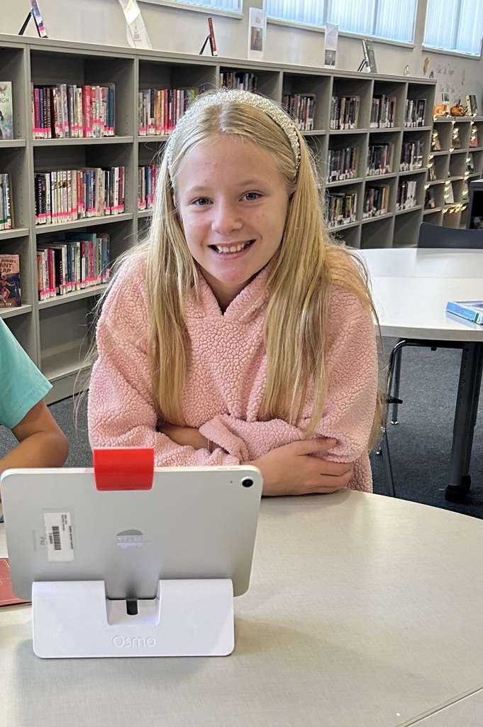 A smiling girl with long blonde hair and a headband sits at a library table in a pink fuzzy hoodie. She is using an iPad in an Osmo stand, with bookshelves visible in the background.