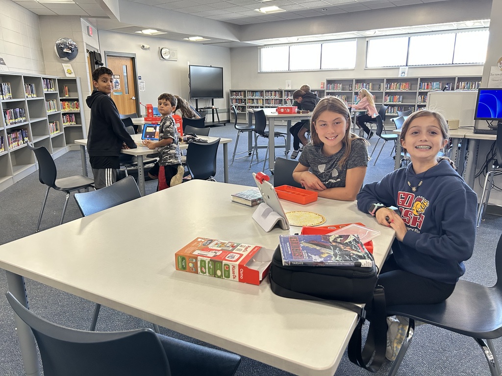 A school library filled with students. In the foreground, two smiling girls are seated at a table with an Osmo game and books. Bookshelves line the background.