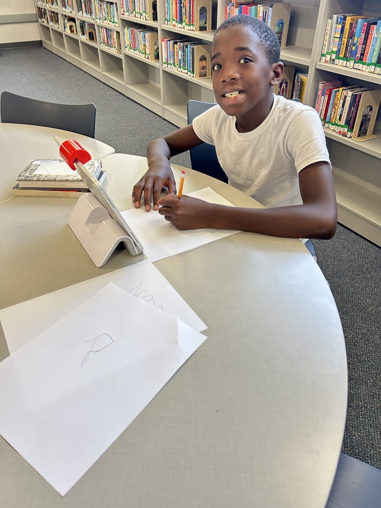 A smiling student sits at a library table, ready to write or draw with a pencil on paper. He is using an Osmo drawing program on an iPad, with bookshelves behind him.