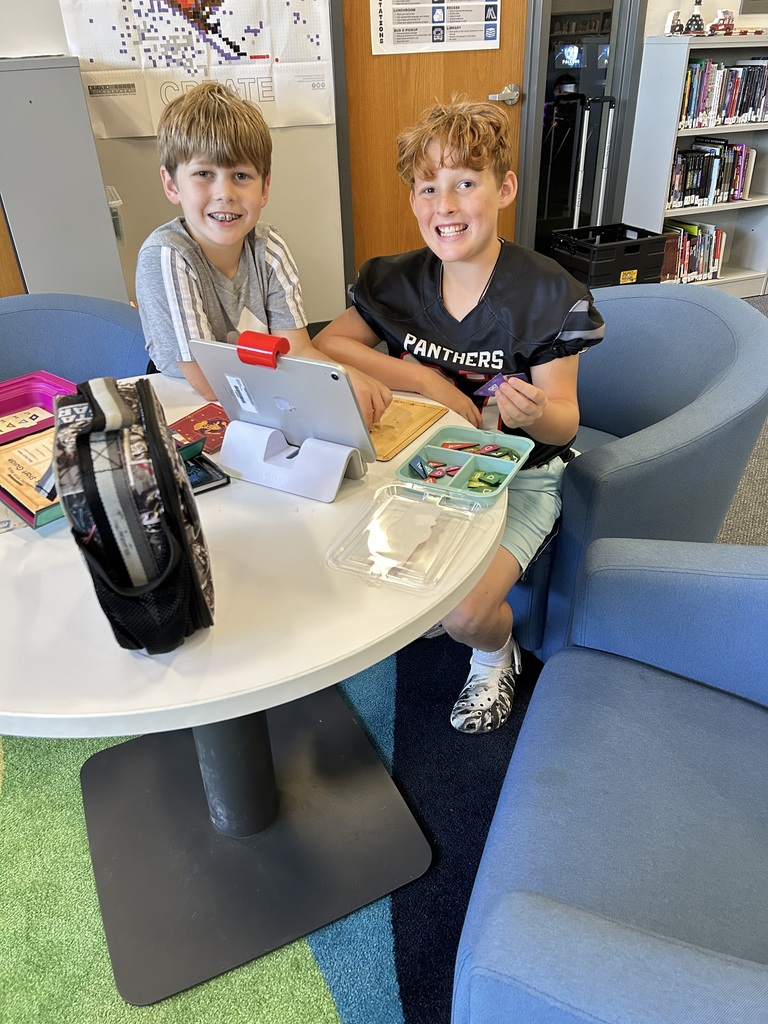 Two smiling students are seated in blue club chairs at a round table. They are interacting with an Osmo learning game on an iPad, using trays of colorful game pieces.