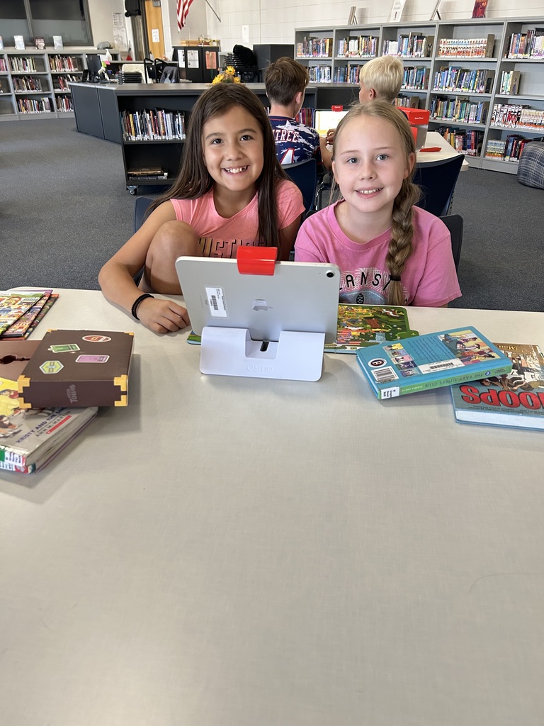 Two smiling girls are seated closely together at a library table with an iPad and several stacks of books. They are smiling warmly at the camera.