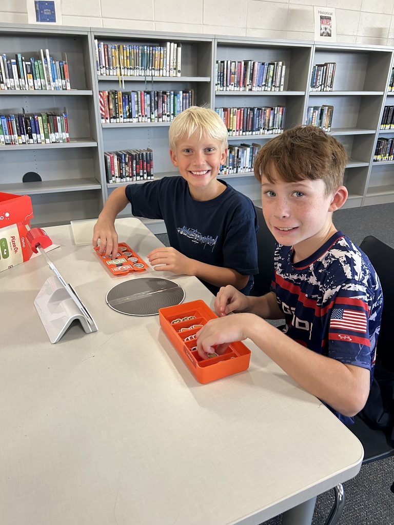 Two smiling boys are at a table in front of library shelves. They are playing an Osmo game, interacting with trays of colorful pieces in front of an iPad.