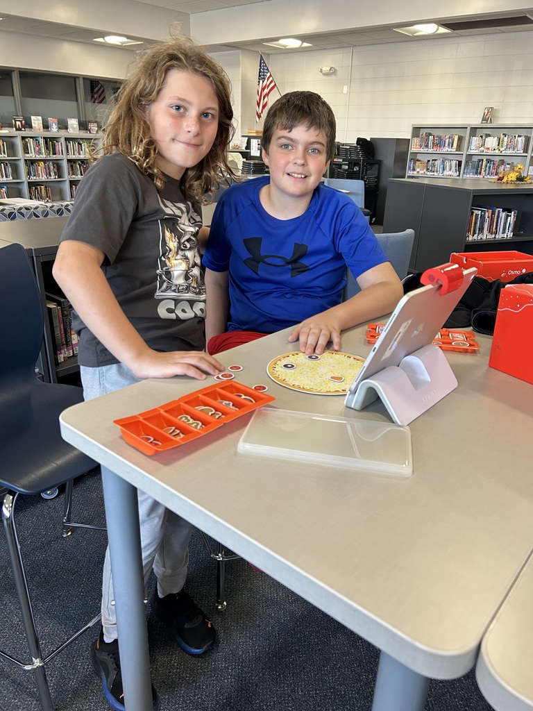 Two happy students pose at a library table, playing with an Osmo Pizza Co. interactive game. There are game pieces, an iPad, and an American flag in the background.