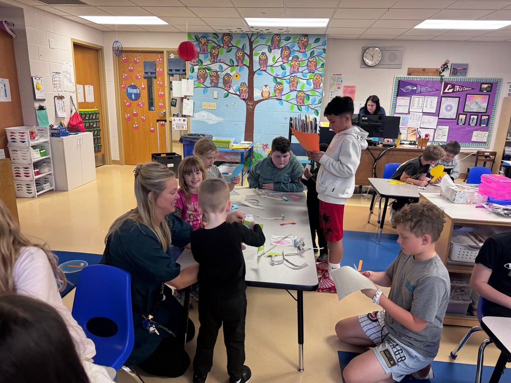 eacher assisting a young student with an art project in a classroom featuring an owl mural and active children.
