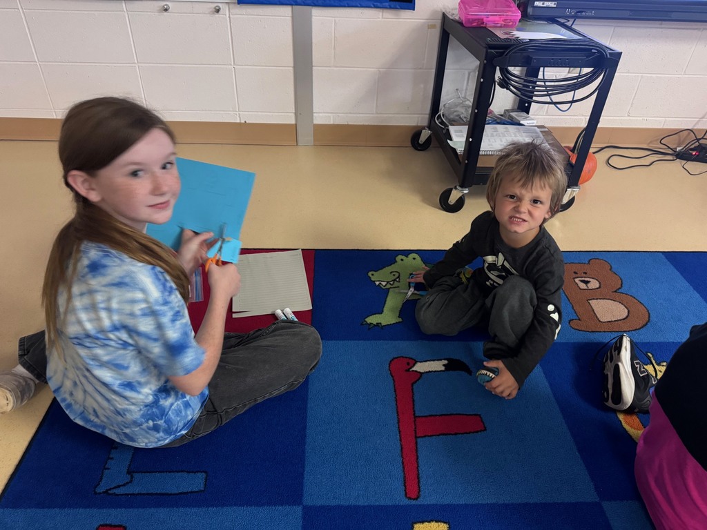 Students sitting on a colorful alphabet rug in a classroom, with one girl cutting blue paper and a boy smiling
