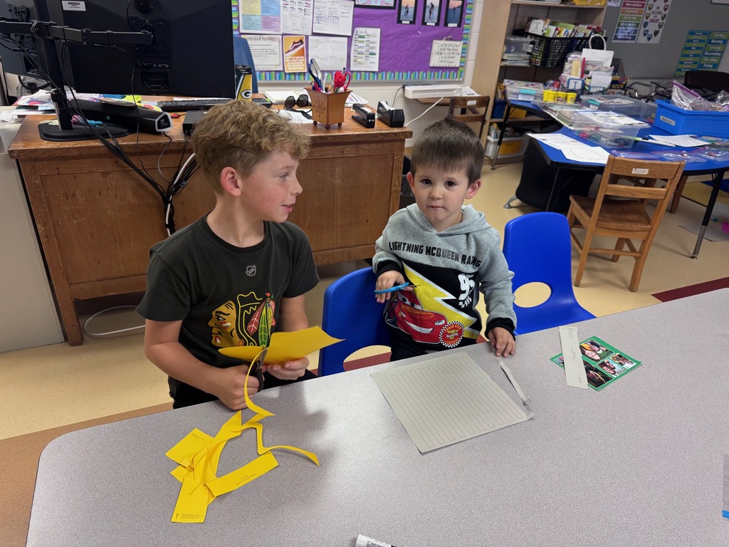 Two young male students sitting at a table in a classroom, focused on cutting yellow construction paper with safety scissors.