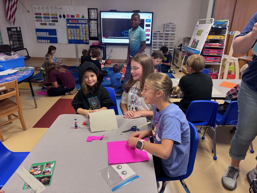 Elementary students engaged in arts and crafts activities at desks and on the floor in a bright, colorful classroom.
