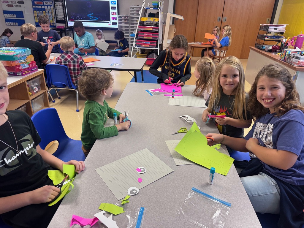 Students smiling and collaborating on hands-on papercraft projects at tables in a lively classroom.