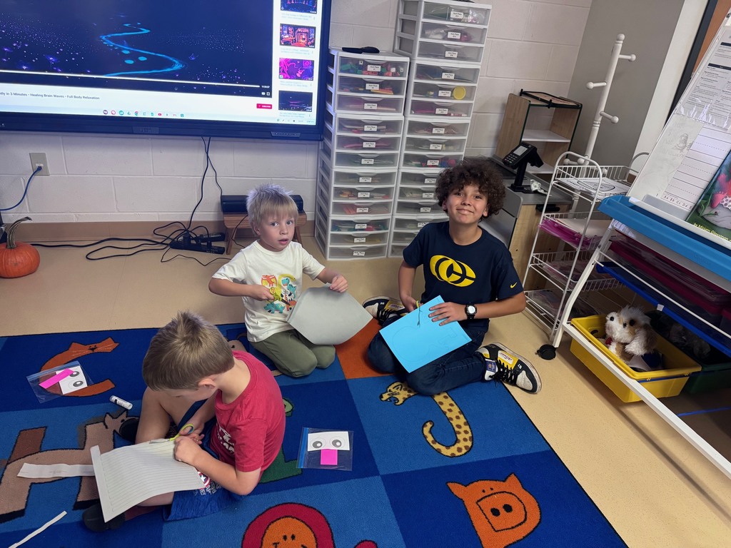 Three students sitting on a colorful carpet in a classroom, working on crafts and cutting paper.