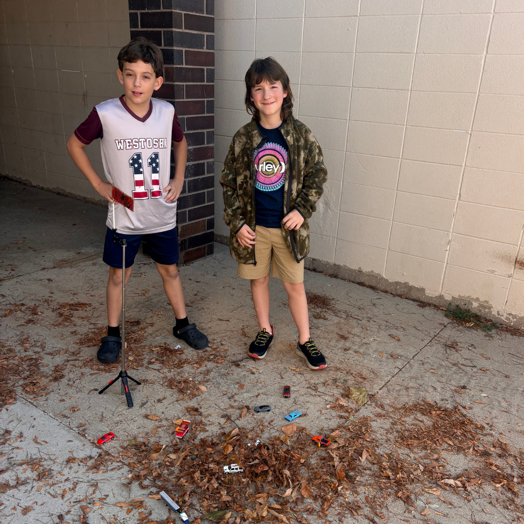 Two young students standing outside near leaves and small toy cars on the ground.