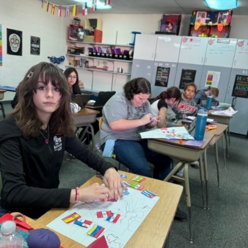 A student arranging colorful country flag cutouts on a map worksheet at her desk in a classroom.