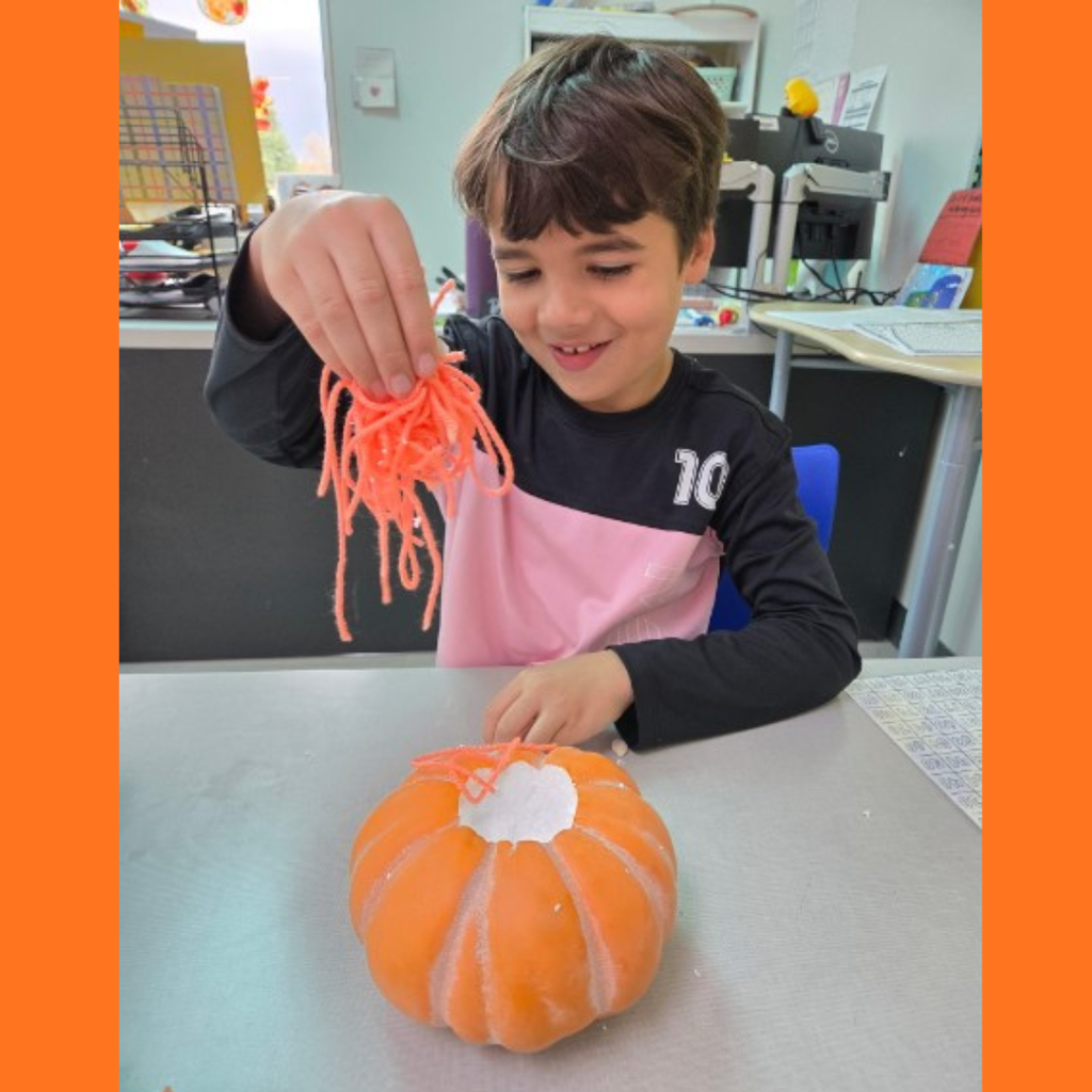 A smiling young male student placing orange yarn inside a craft pumpkin during a classroom lesson.