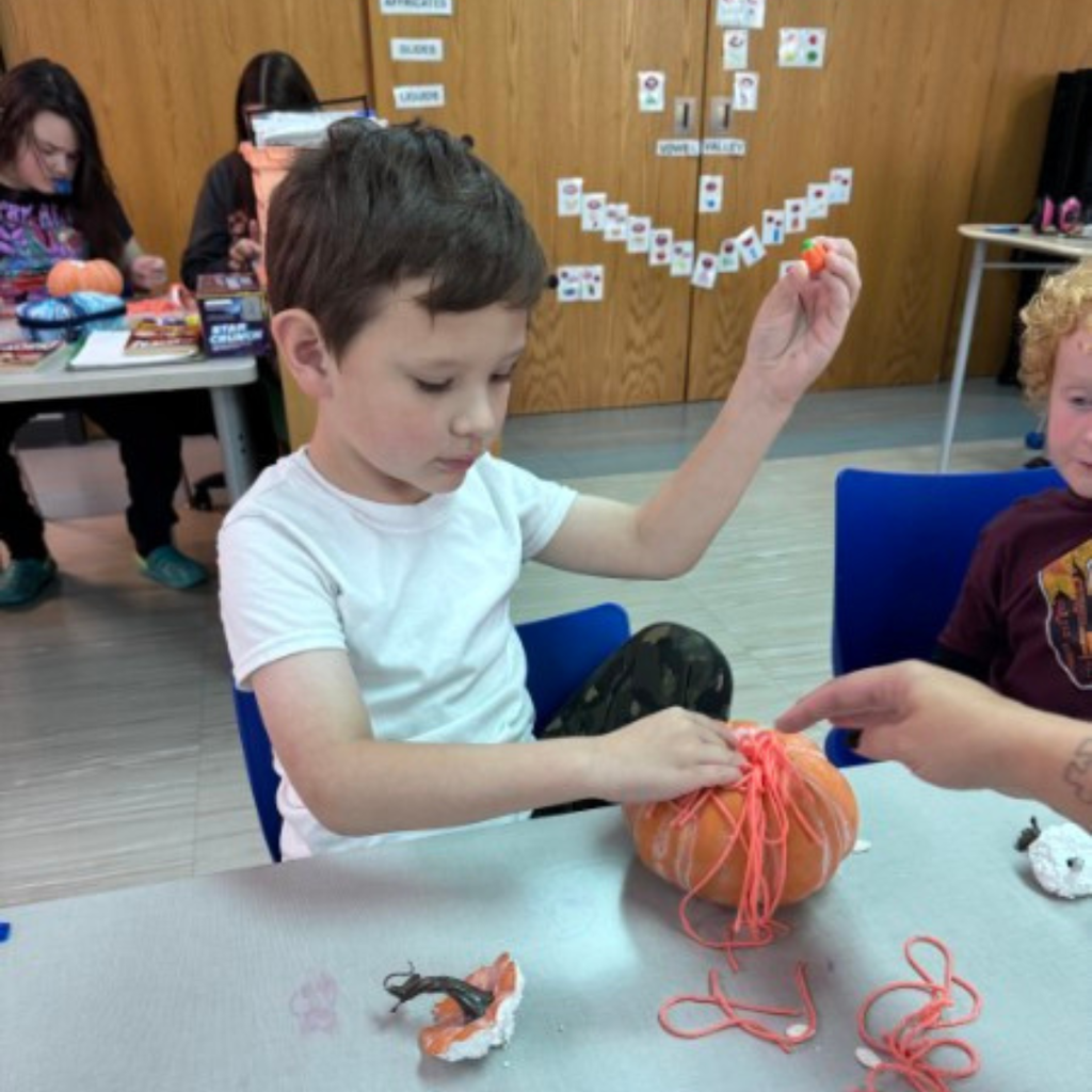 A young student focusing on adding orange yarn strands to a craft pumpkin during a hands-on class activity.