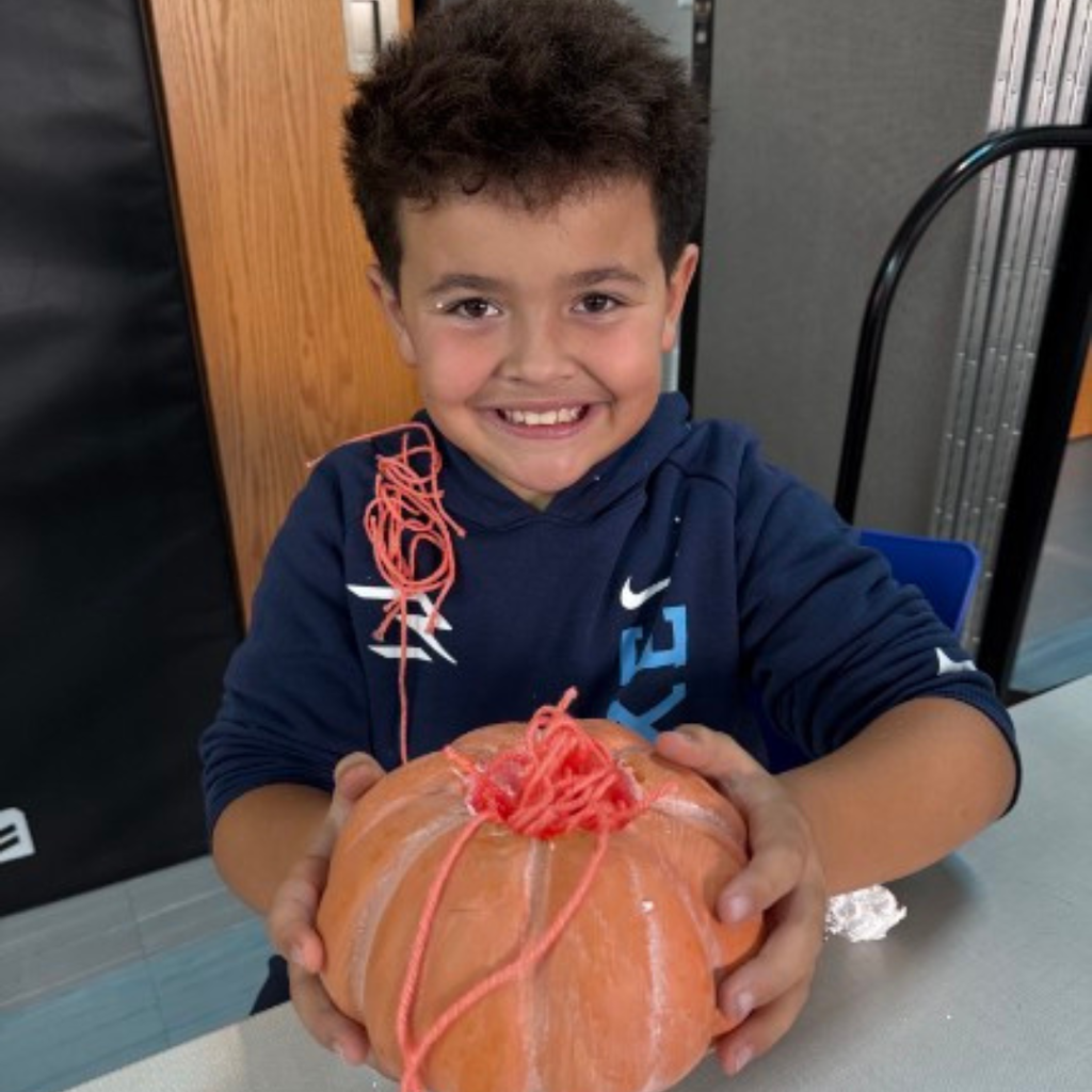 A young student smiling as he proudly presents his finished craft pumpkin filled with orange yarn.