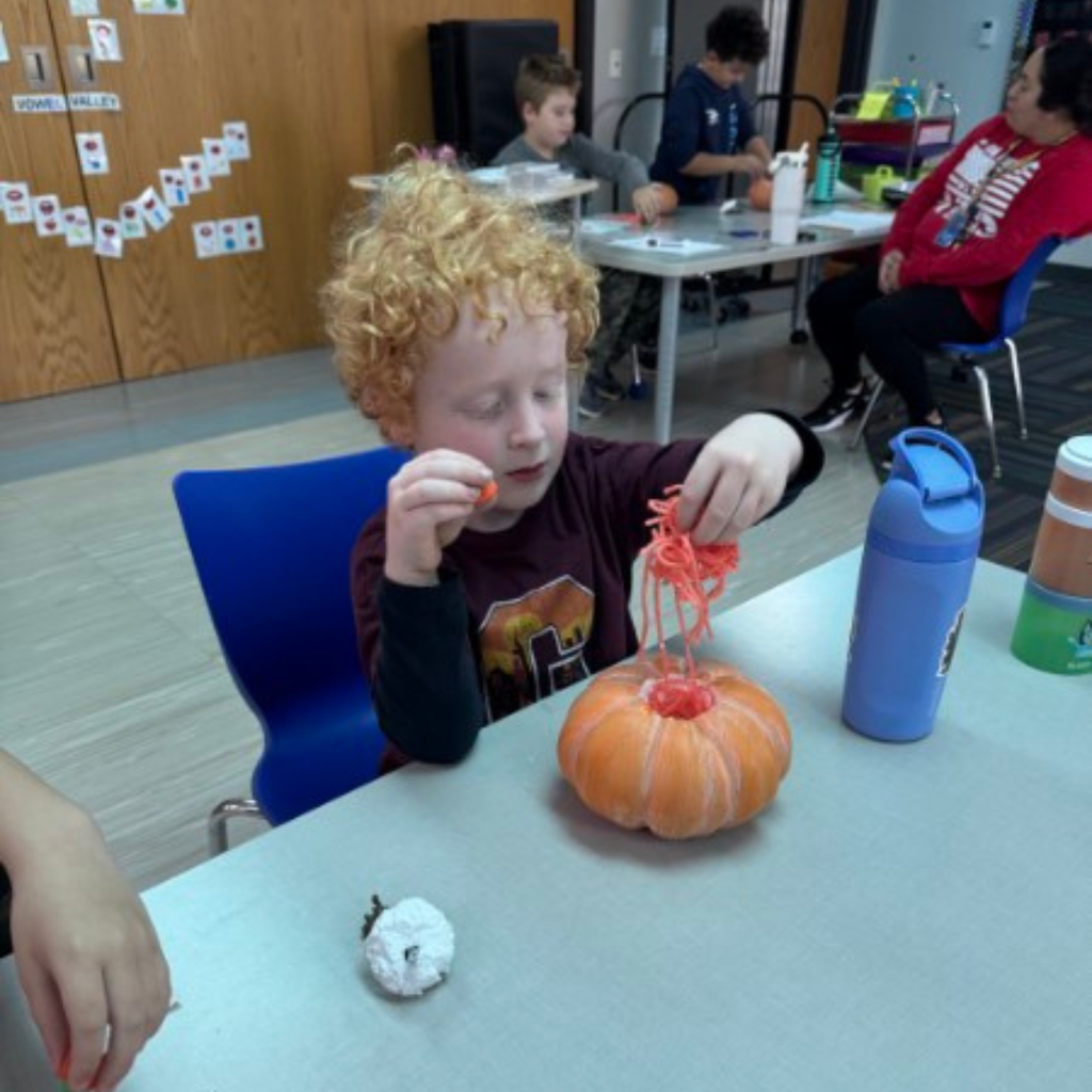 A young student with curly hair placing orange yarn into a craft pumpkin at a classroom table.