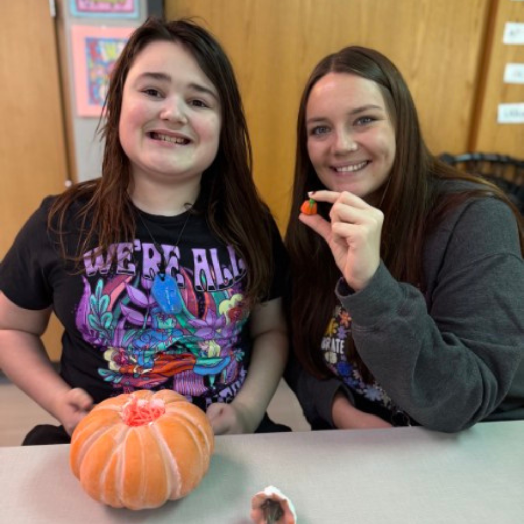 A student and teacher smiling together at a classroom table with a pumpkin craft activity.