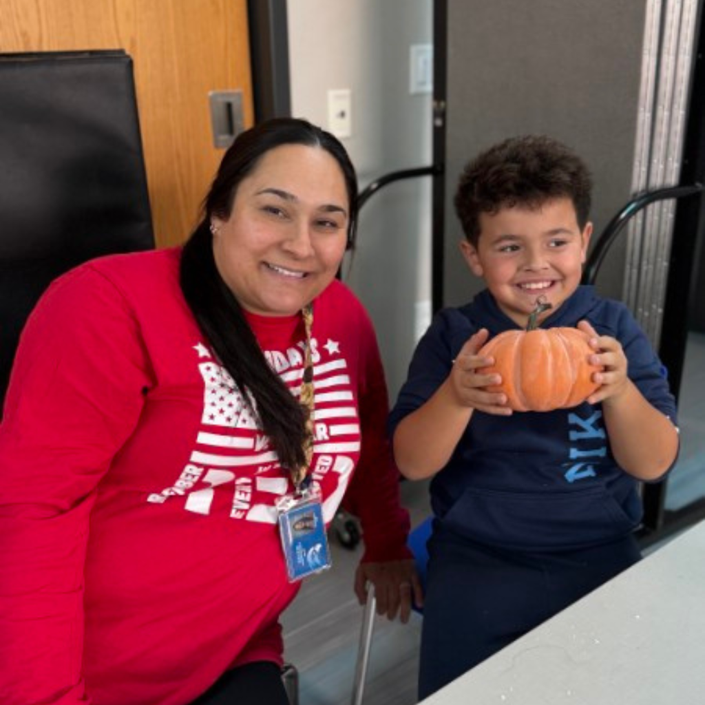 A teacher and a smiling young student posing together at a table while holding a completed craft pumpkin.
