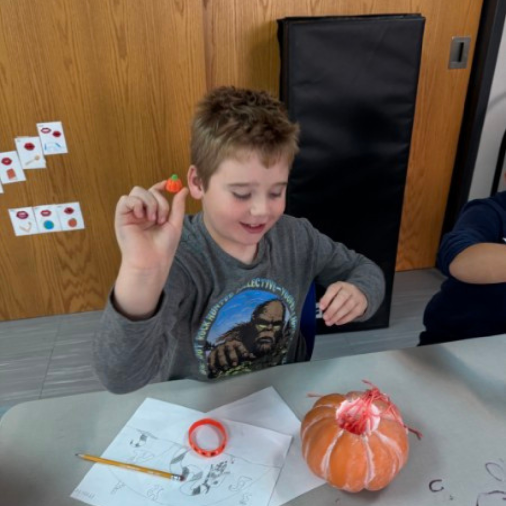 A student smiling as he holds up a tiny plastic pumpkin piece next to his pumpkin craft at his desk.