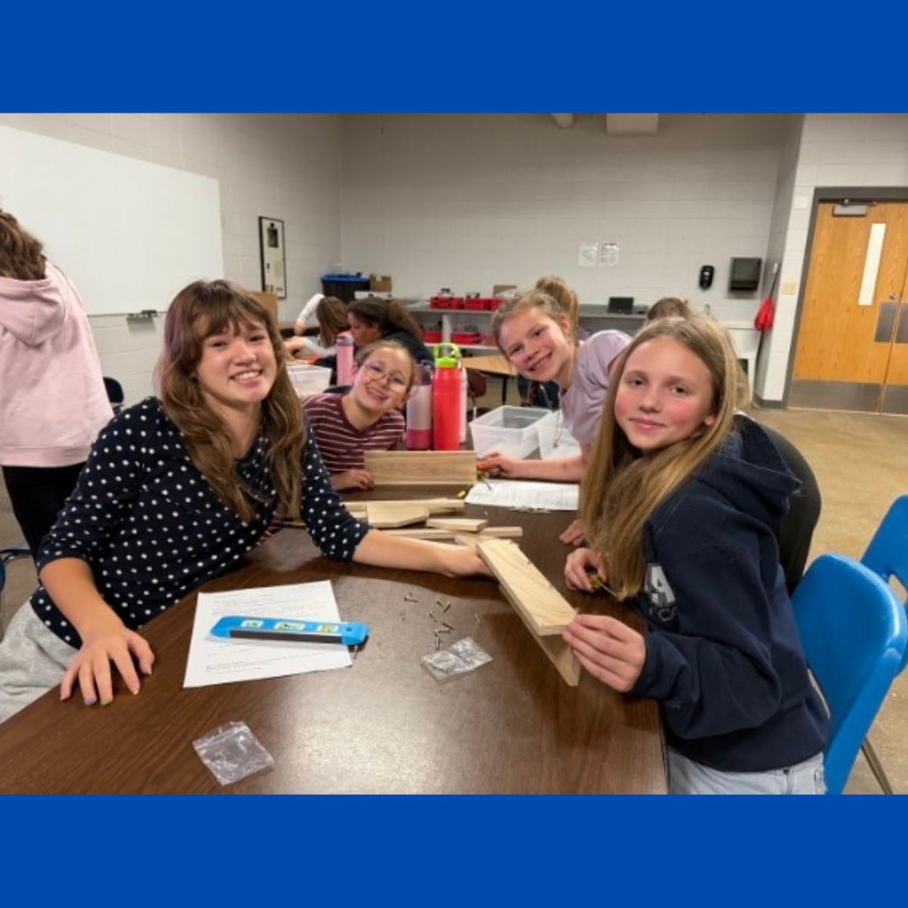 social (70).jpg: Four female students smiling together at a wooden table while working on a woodworking project with screws and tools.