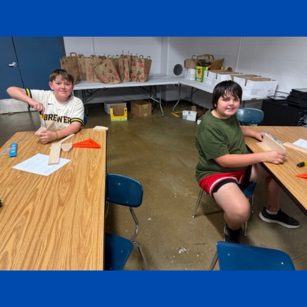 Two students sitting at classroom tables using screwdrivers to assemble wooden building projects.