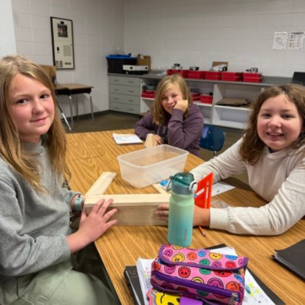Three female students seated around a wooden desk smiling as they work together on a building project.