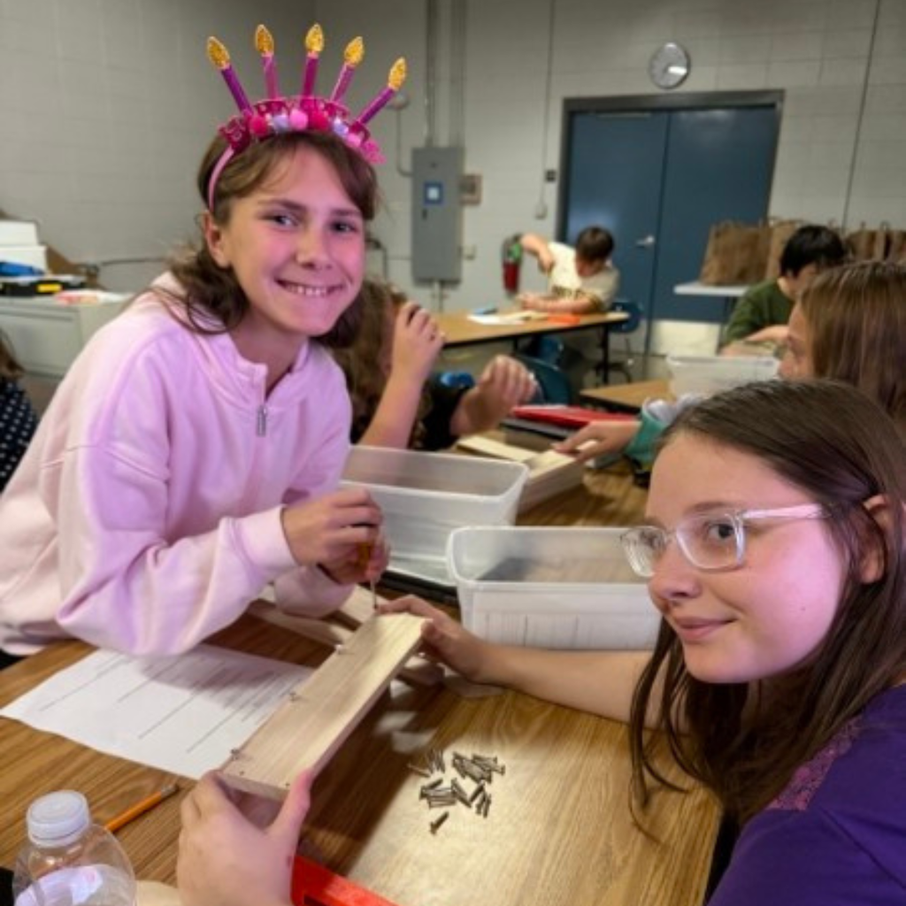 Two students smiling while working together on a wooden assembly project with screws at a classroom table.