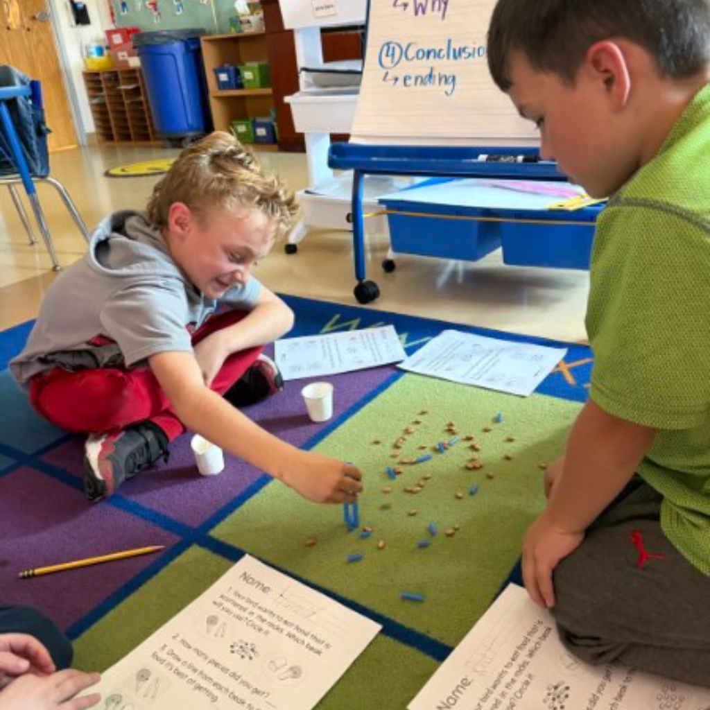 Four young students working together on a rug in front of an anchor chart easel during a classroom lesson.
