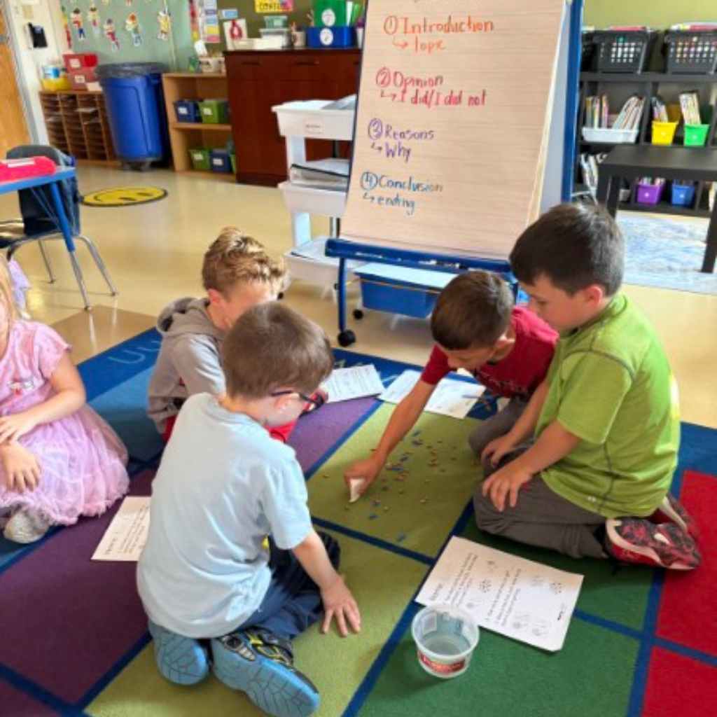 Two young students working together on a classroom floor activity using plastic tweezers and worksheets.