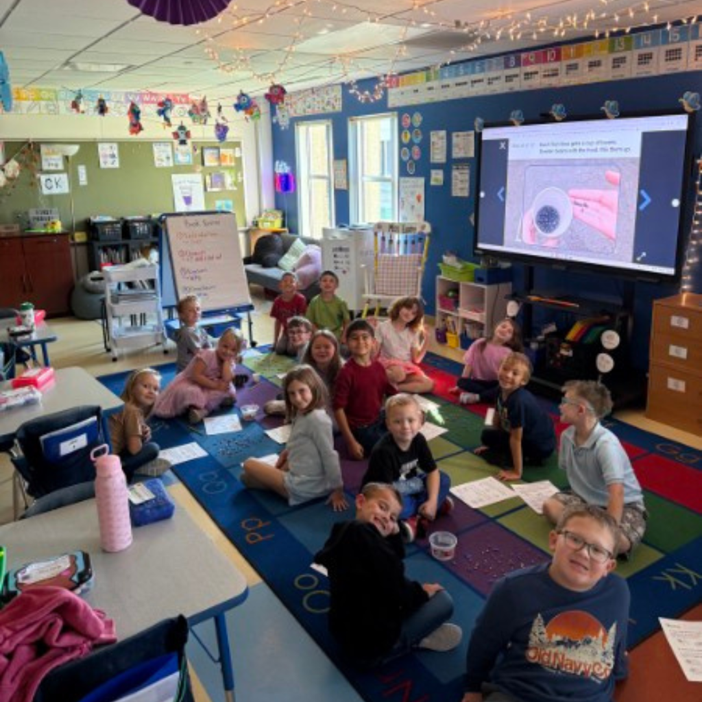 Students sitting on a carpeted classroom floor using tweezers to gather small items for a science activity.